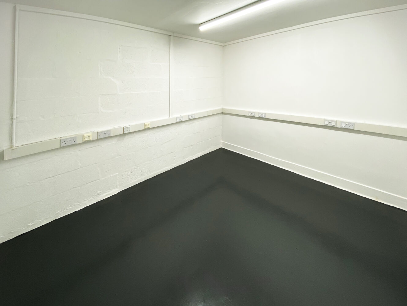 Empty white-walled room with a black floor, fluorescent ceiling light, and electrical outlets along the walls.