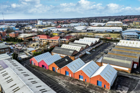 Aerial view of an industrial area with rows of warehouses, some painted red, surrounded by parking lots and scattered buildings under a partly cloudy sky.