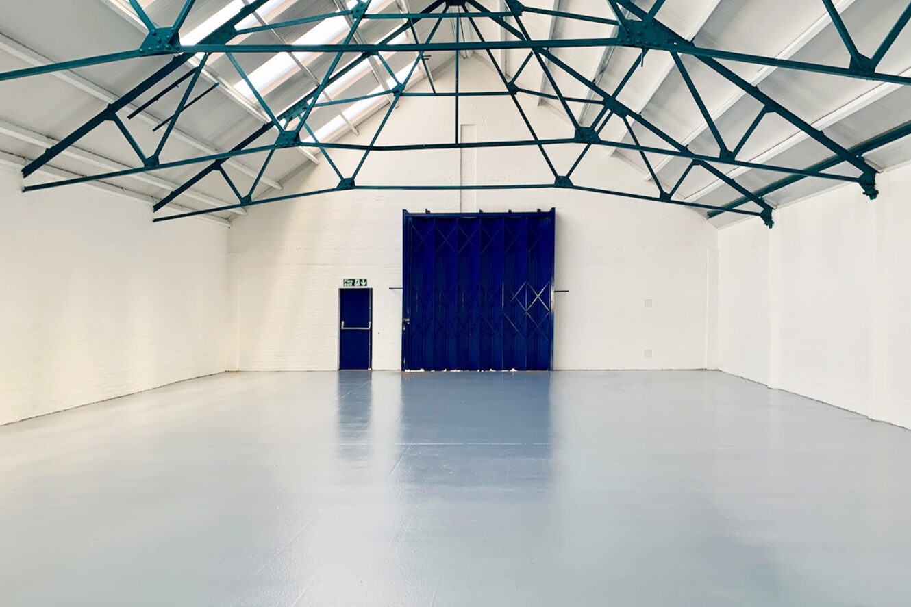 An empty industrial warehouse with white walls, a shiny gray floor, steel roof beams, a blue door, and a large blue folding gate.