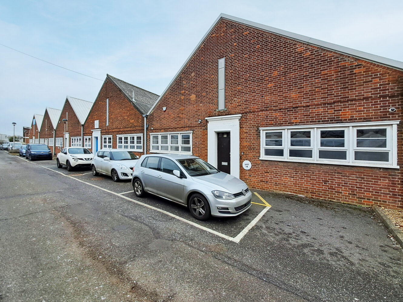 A row of brick industrial buildings with sloped roofs and parked cars in front along a paved street on a cloudy day.