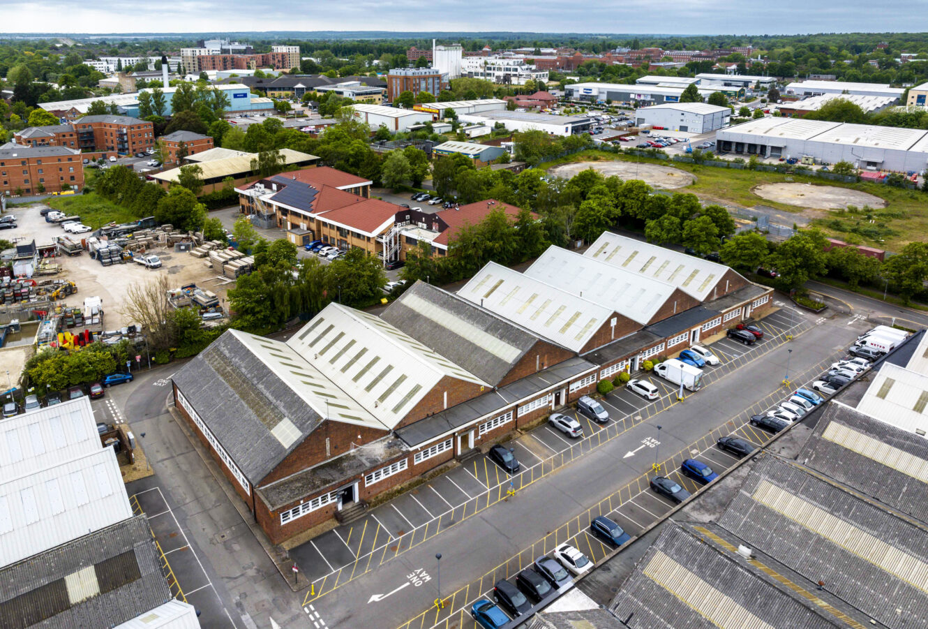Aerial view of industrial warehouses with parking lot, surrounded by trees and other buildings in an urban area.