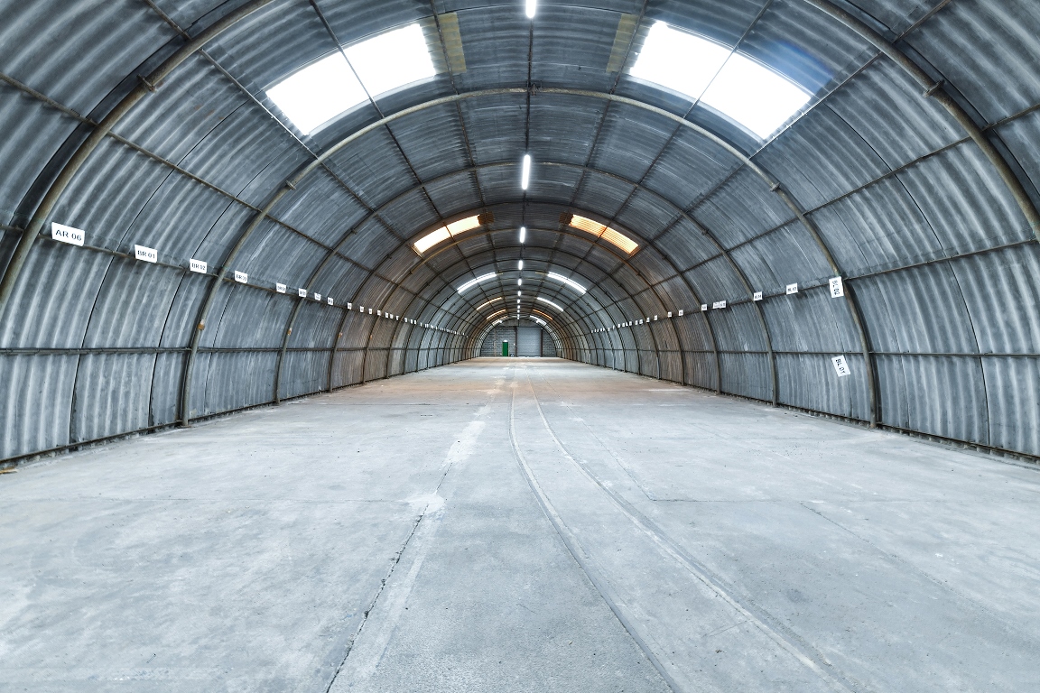 Empty arched warehouse with corrugated metal walls and ceiling, skylights, and concrete floor, viewed from the center looking toward the far end.
