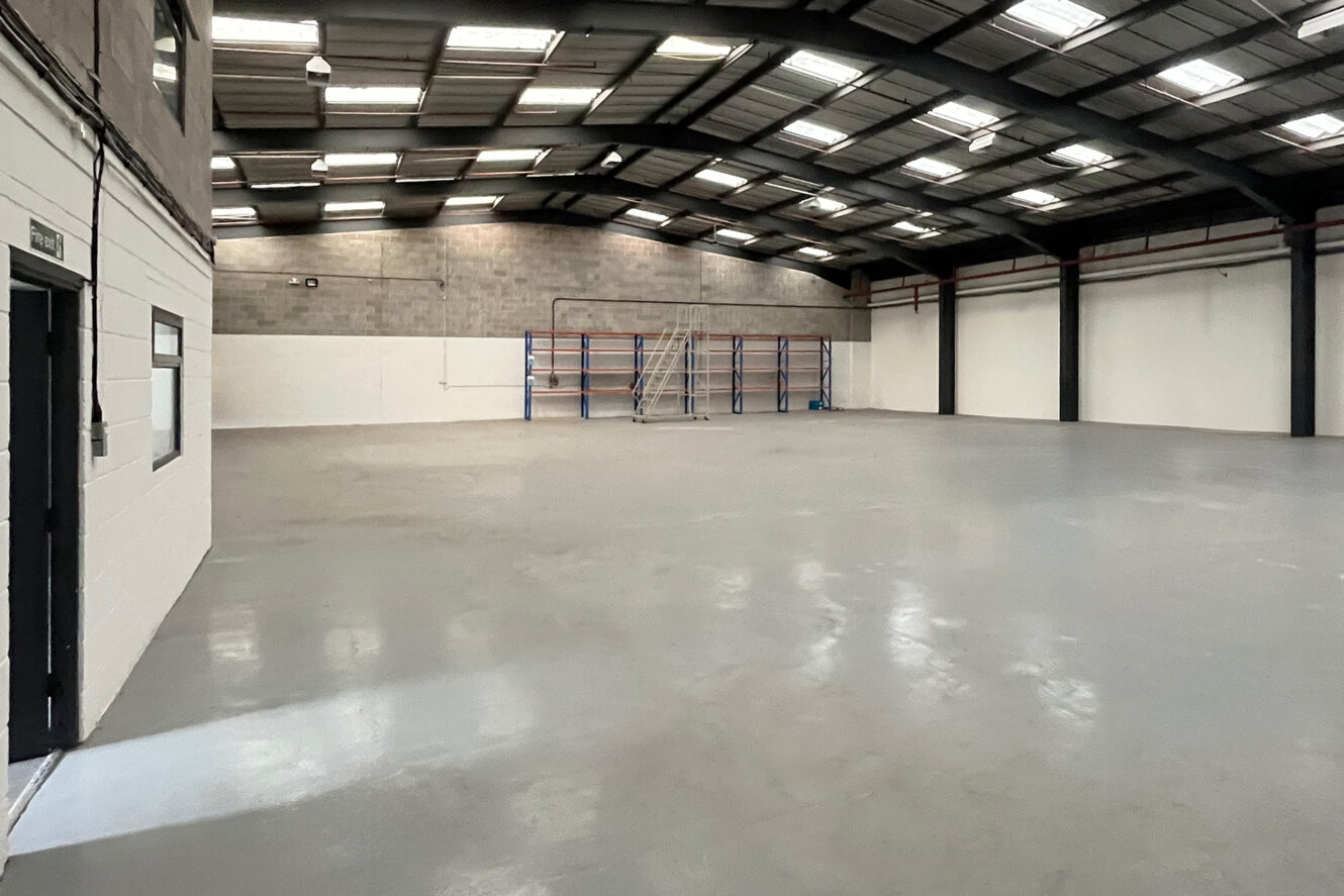 Interior of an empty industrial warehouse with concrete floor, metal roof, white walls, and a storage rack with a ladder against the back wall.