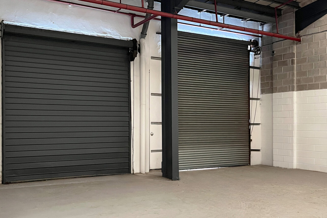 Two large industrial roller doors, one black and closed, the other metallic and partially open, in an empty warehouse with concrete floor and brick walls.
