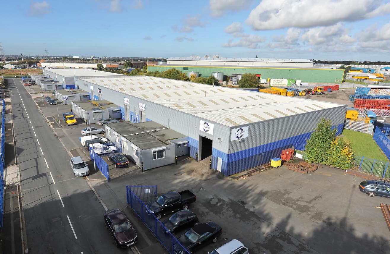 Industrial warehouse complex with blue and white buildings, parked cars, and office units along a paved road under a partly cloudy sky.