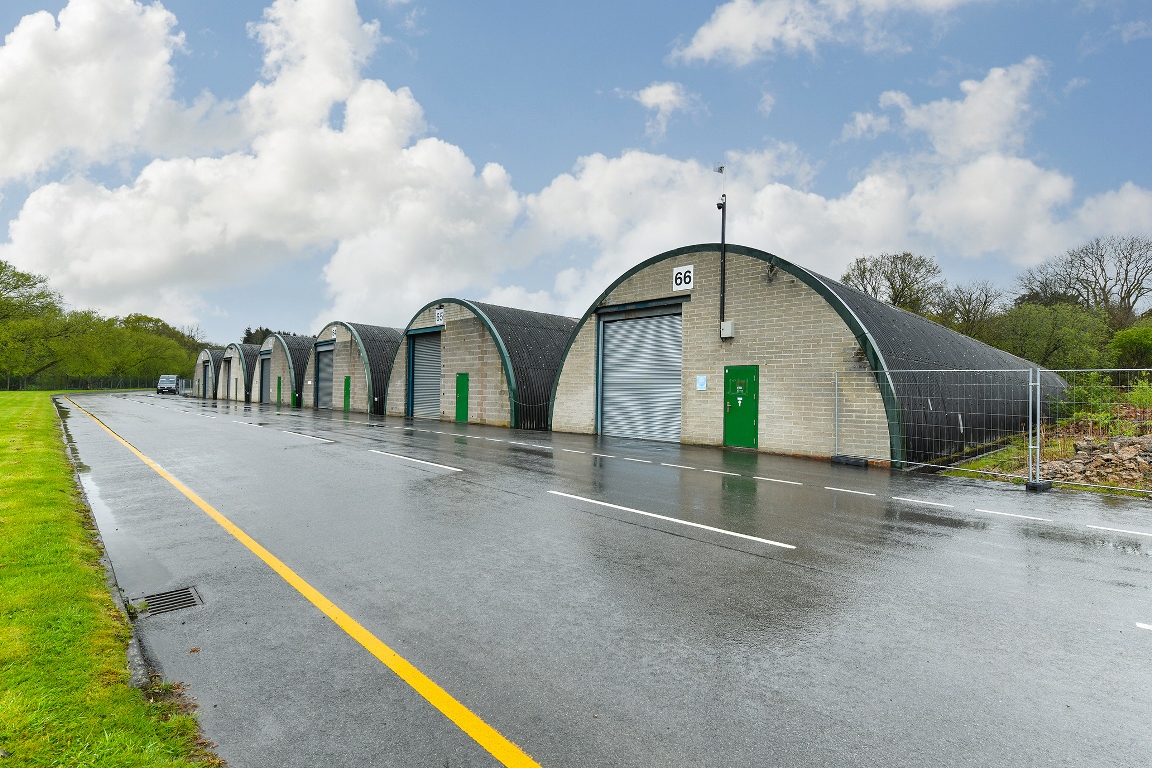 A row of arched-roof storage units with green doors and one metal roller door, situated along a wet road under a cloudy sky.