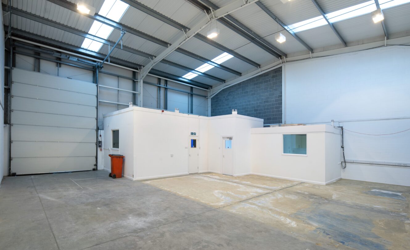 Interior of an empty industrial warehouse with high ceilings, exposed beams, a roller shutter door, white partitioned office rooms, and a red trash bin.