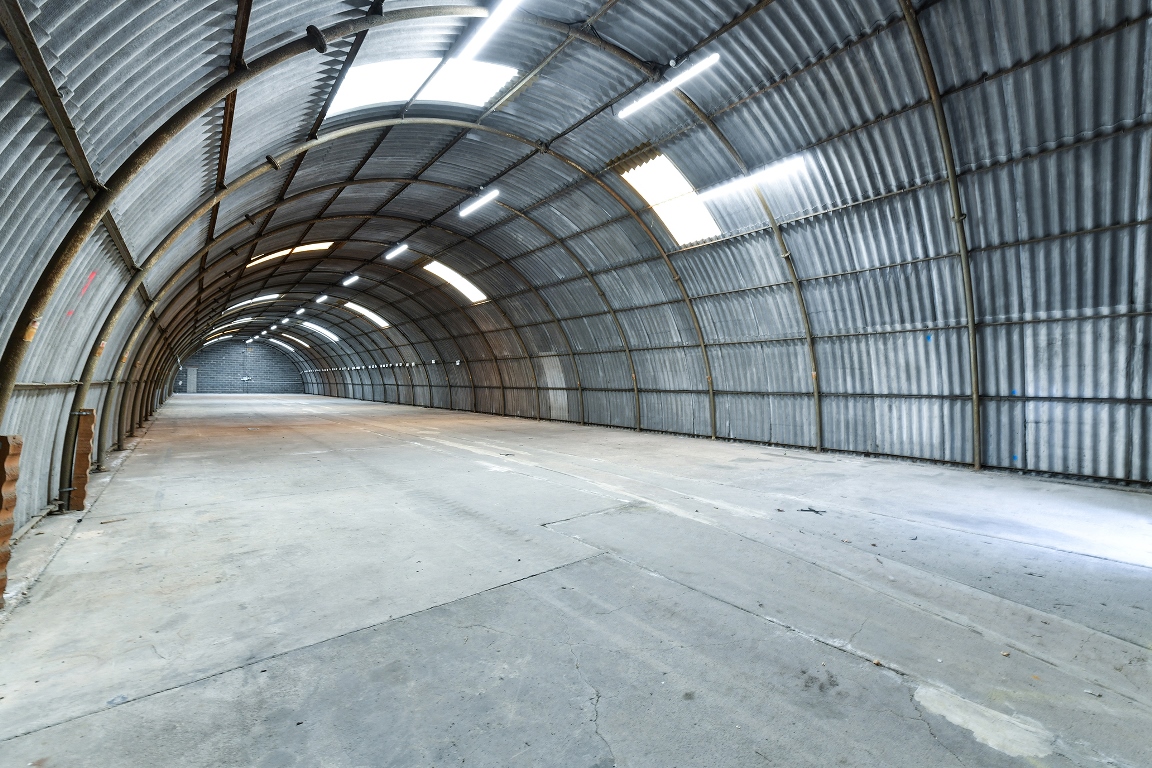 Wide-angle view of an empty, arched metal warehouse with a concrete floor and overhead fluorescent lighting.
