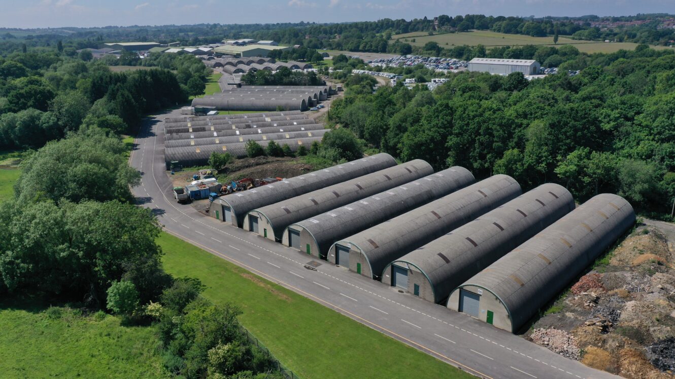 Aerial view of several long, curved-roof warehouse buildings lined up along a road, surrounded by trees and open fields.