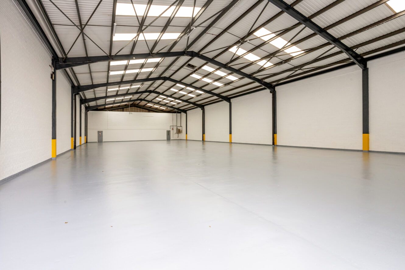 Spacious empty warehouse with white walls, high ceiling, exposed beams, and large skylights allowing natural light. Yellow safety markers are on the lower wall sections.