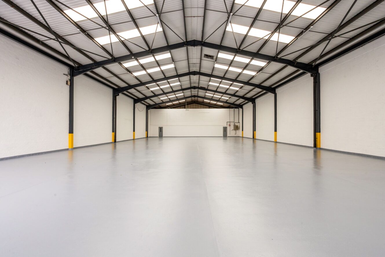 Interior of an empty industrial warehouse with white walls, polished gray floor, high ceiling, and exposed steel beams. Bright overhead lights and skylights provide illumination.