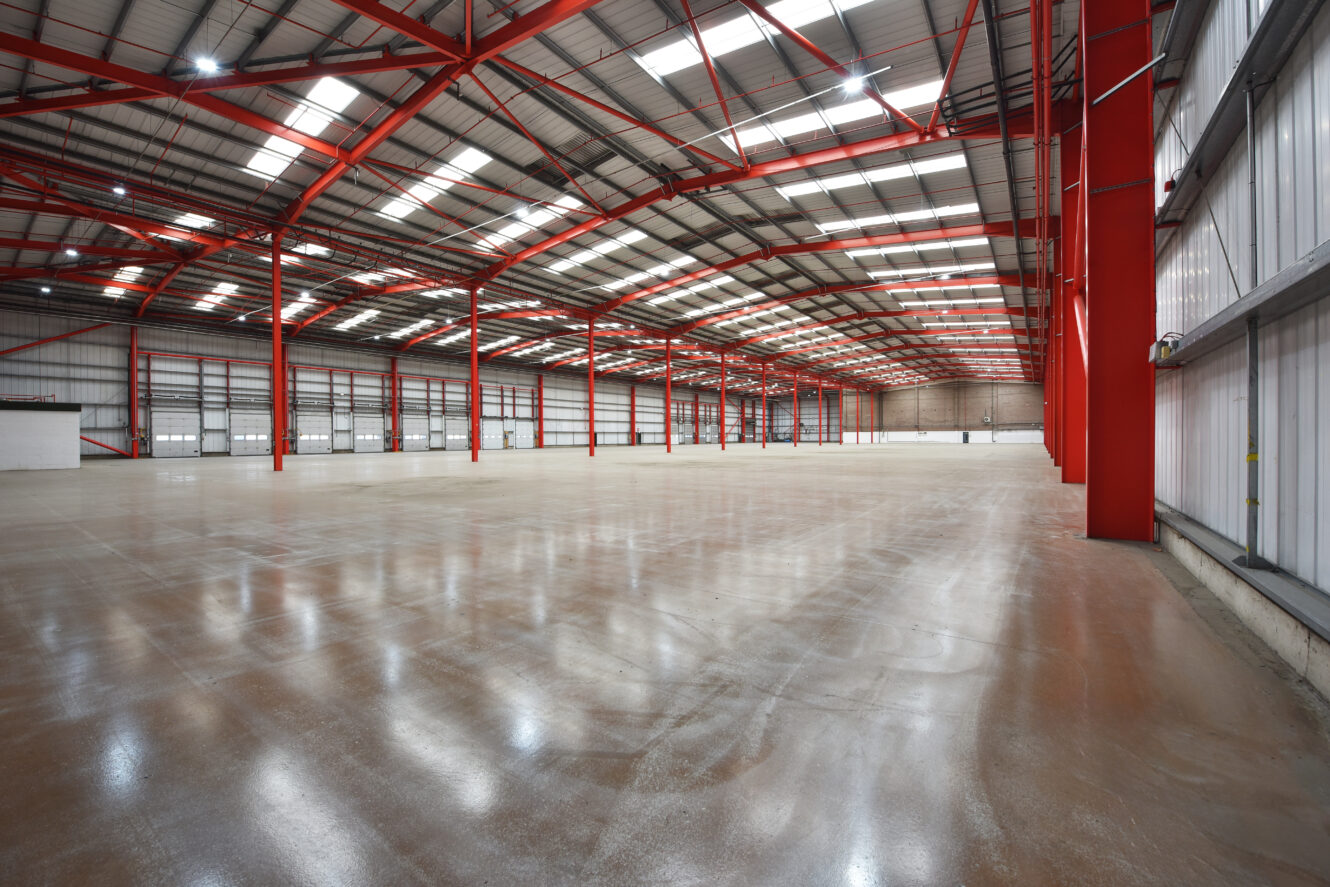 Interior of a large, empty warehouse with red steel beams, high ceiling, polished concrete floor, and bright overhead lighting.