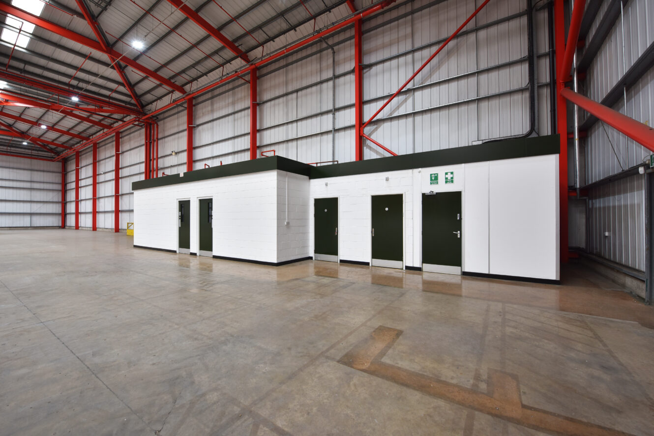 Large empty warehouse interior with high ceilings, exposed beams, and a small white walled structure with several green doors in the center.