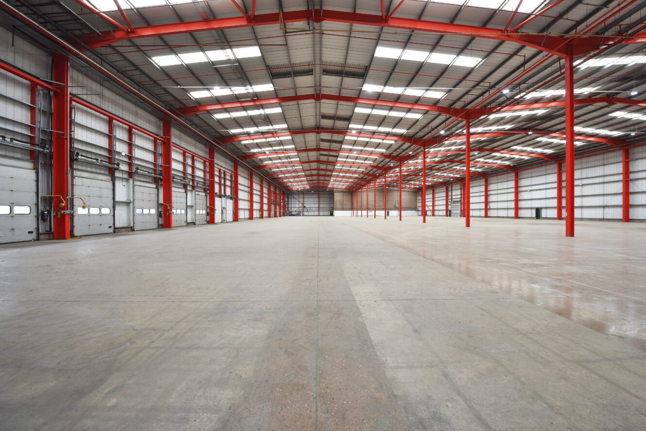 Large, empty warehouse interior with high ceilings, red steel beams, and multiple loading dock doors along the left wall. Concrete floor extends throughout the space.