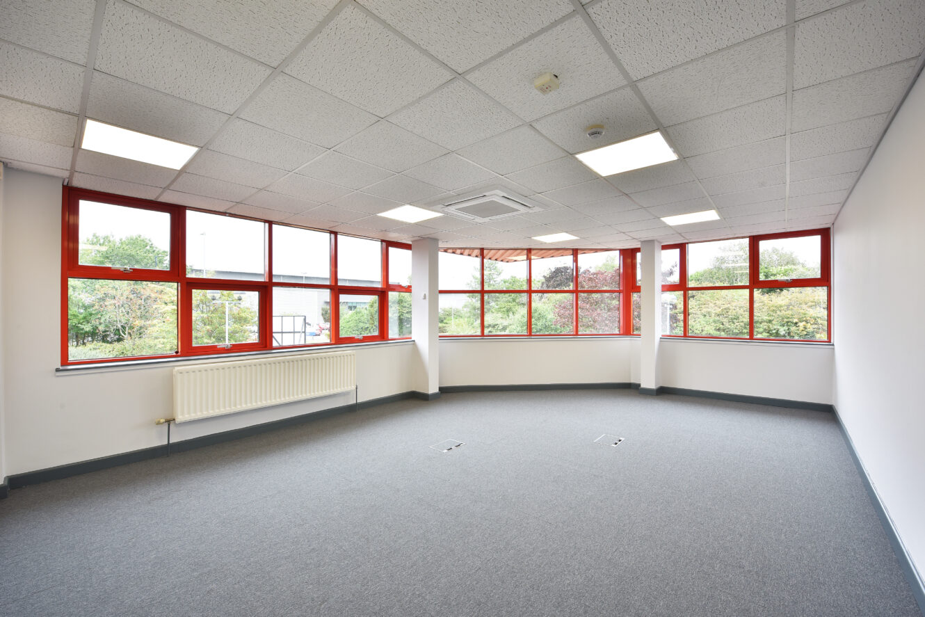 Empty office room with red-framed windows, white walls, gray carpet, ceiling lights, and a radiator beneath the windows, overlooking greenery outside.