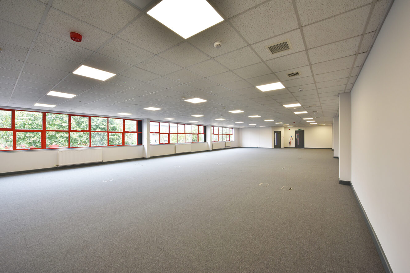 Large empty office space with gray carpet, white walls, tiled ceiling with fluorescent lights, red-framed windows, and a view of trees outside.