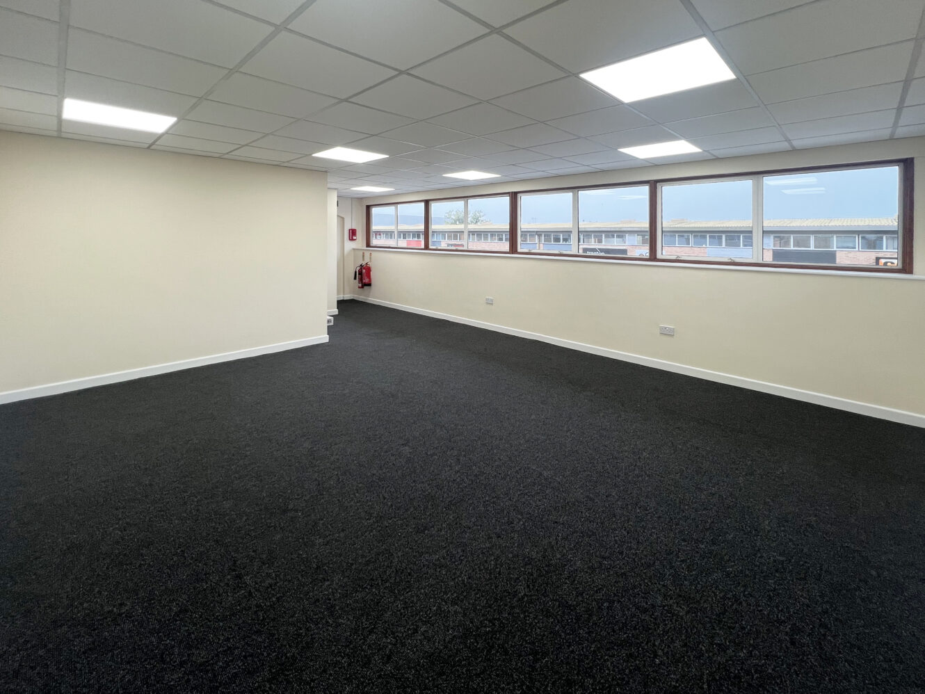 An empty office room with black carpet, cream-colored walls, a ceiling with fluorescent lights, and windows along one side showing a view of nearby buildings.