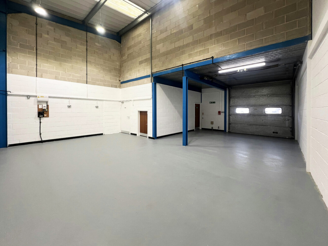 Interior of a clean, empty industrial warehouse unit with painted concrete floor, high ceiling, roller shutter door, and exposed block walls.