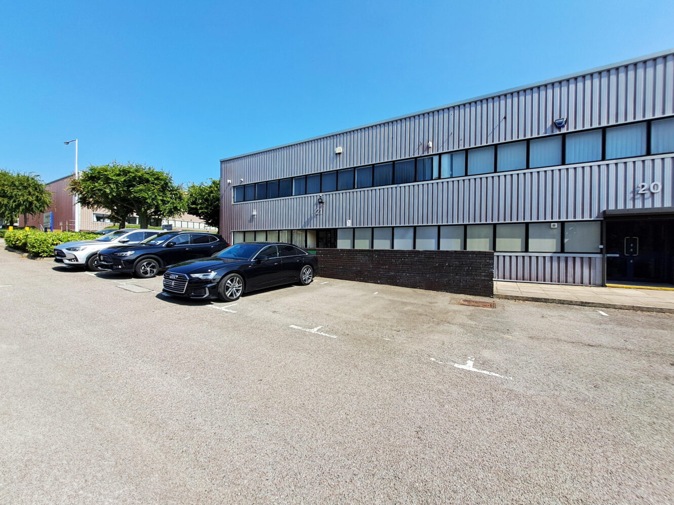 Four cars are parked outside a two-story industrial building with a gray metal exterior on a clear, sunny day.