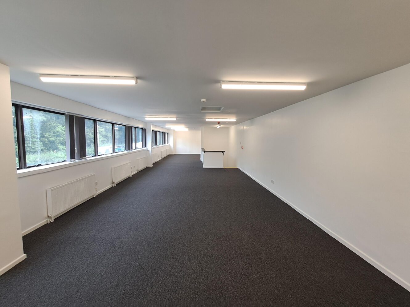 Large, empty office space with white walls, carpeted floor, ceiling lights, and windows along one side. A small stairwell is visible at the far end.