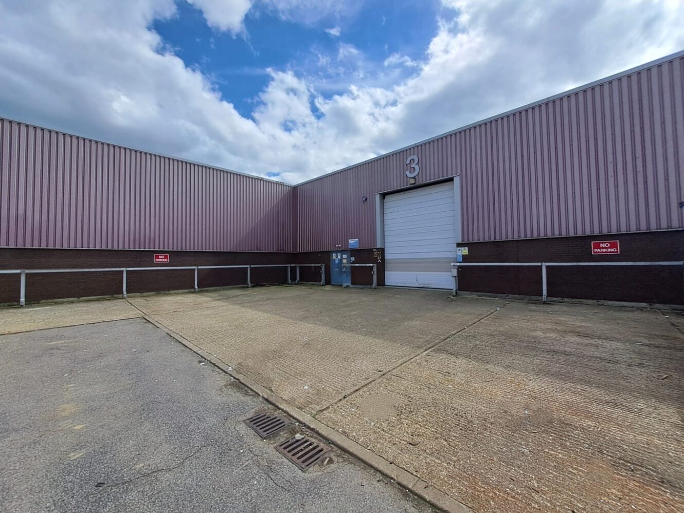A large industrial warehouse with a closed white roller door labeled 3, surrounded by a concrete yard under a partly cloudy sky.