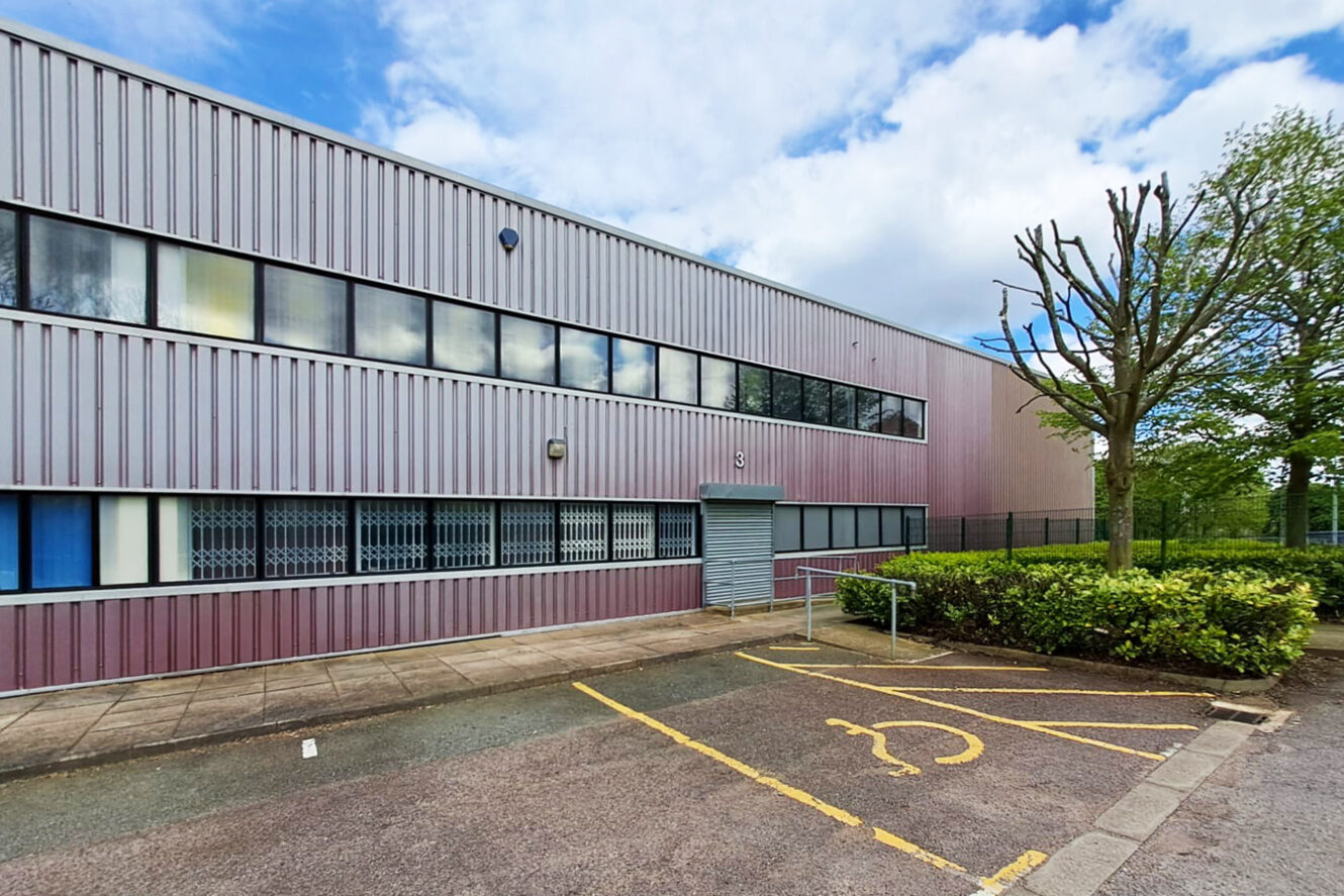 A modern, two-story industrial building with metal siding, large windows, and an accessible parking space in front, bordered by hedges and a tree.