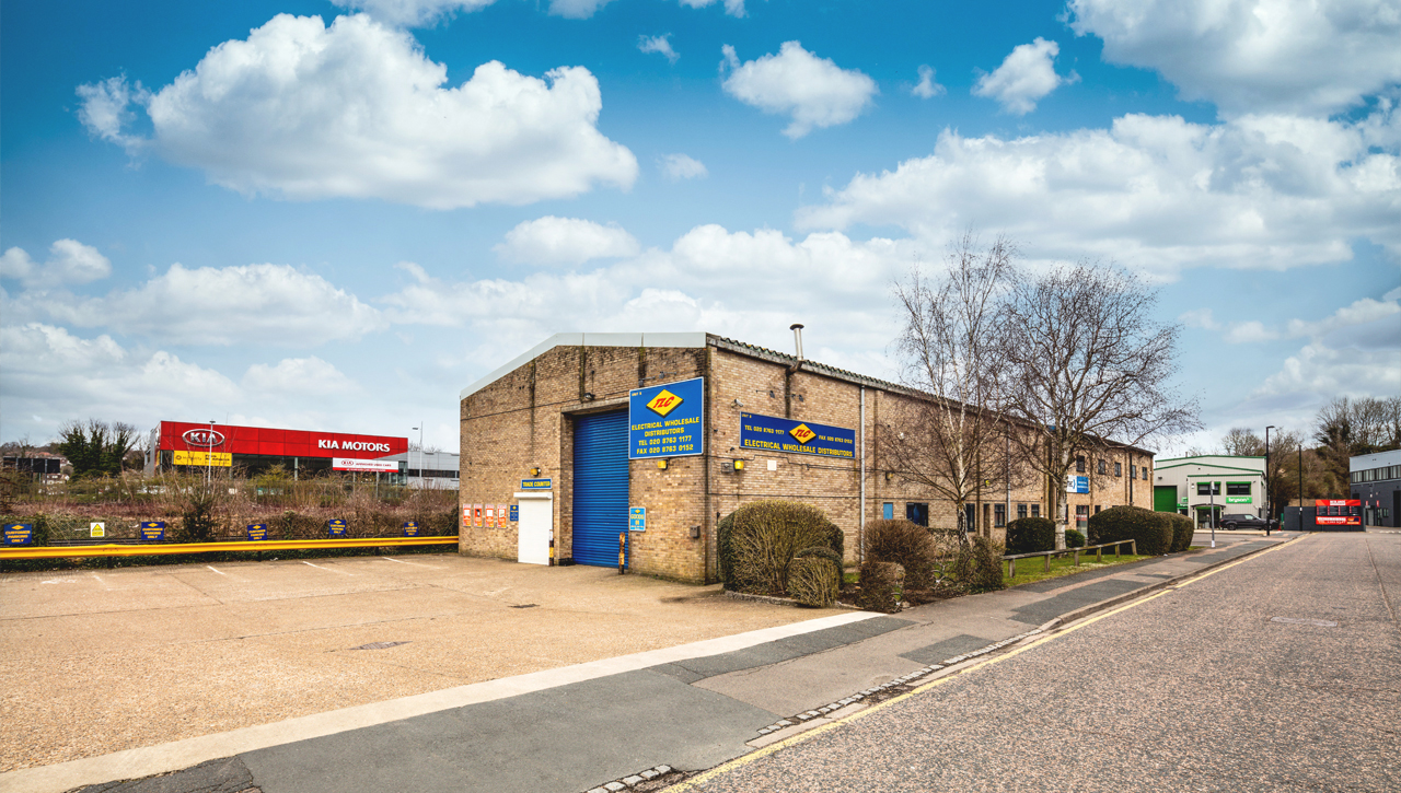 A large industrial warehouse with blue doors and signage sits beside a quiet street on a clear day, with a Kia Motors billboard visible in the background.