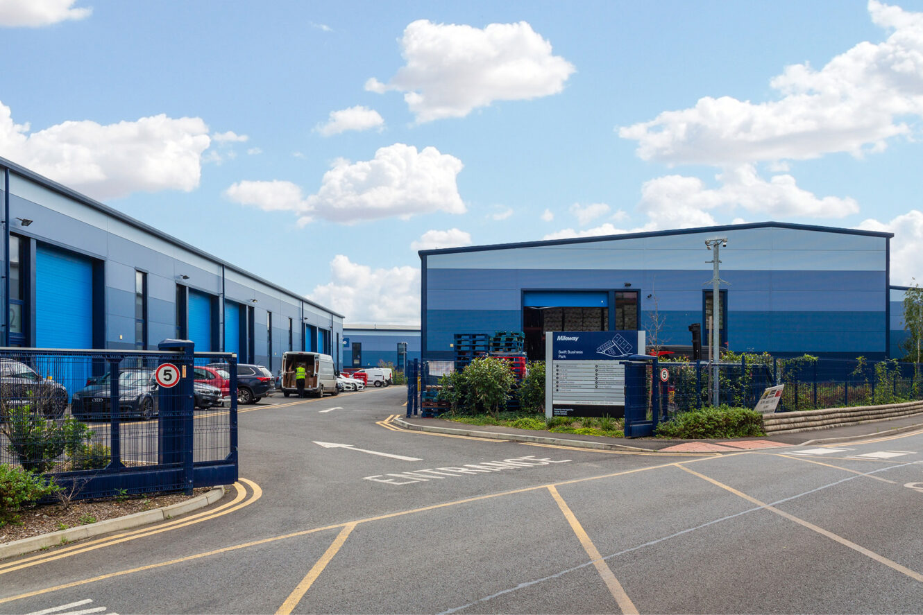 A business park with blue industrial buildings, multiple parked cars, and a sign near the entrance on a partly cloudy day.