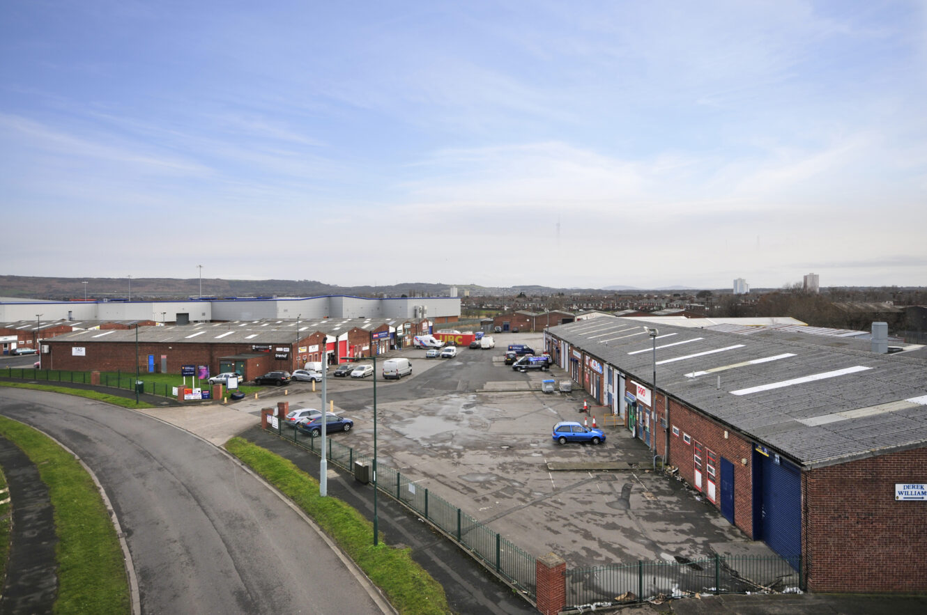 Industrial estate with multiple red-brick warehouses, parked vehicles, and a wide access road under a mostly clear sky. Distant hills and city buildings are visible in the background.