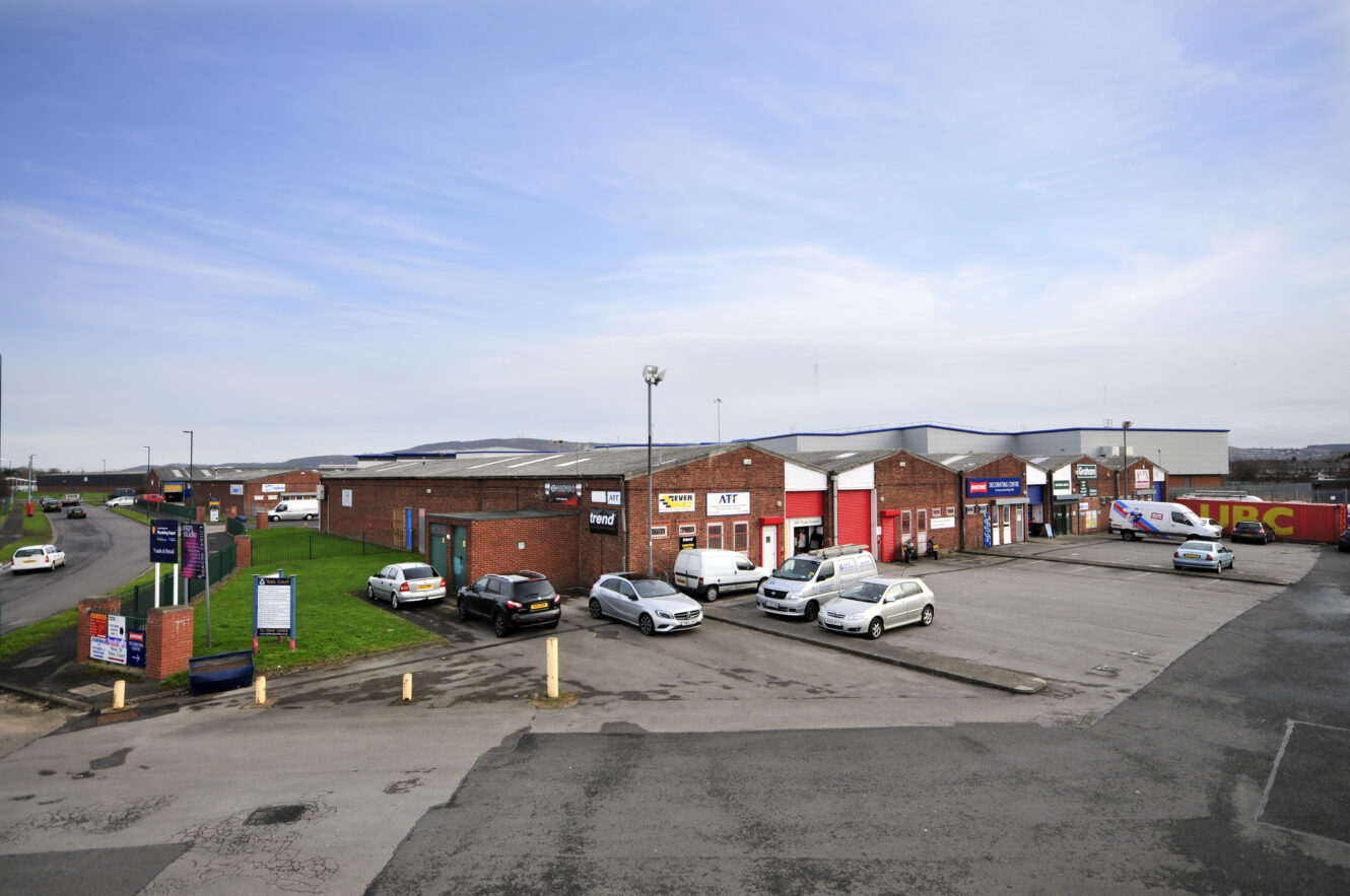 A small industrial estate with brick warehouse units, several parked cars, company signs on buildings, and a mostly empty parking lot under a cloudy sky.