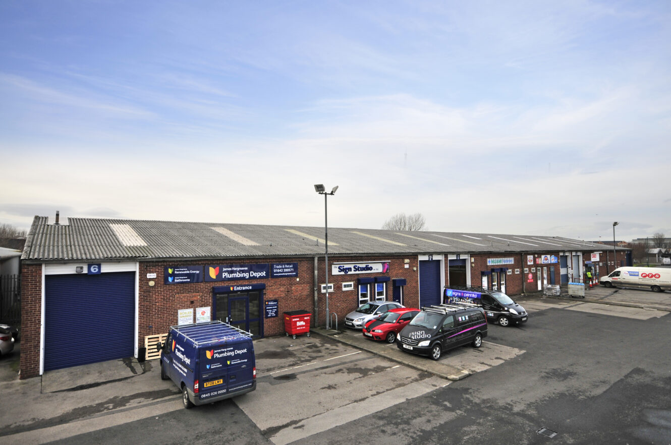 A row of industrial units with businesses, parked cars, and vans in front; signage indicates a plumbing depot and a sign studio.