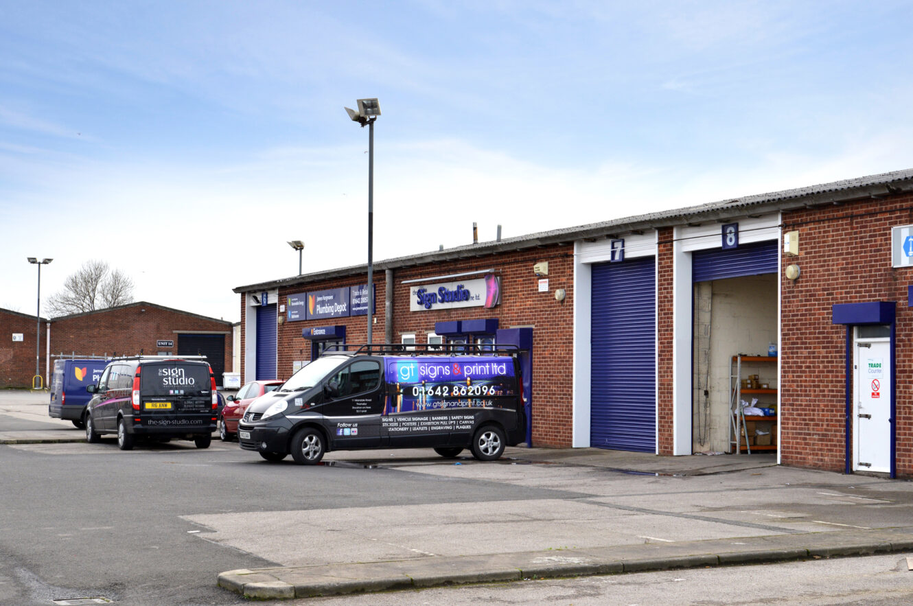 A small industrial estate with brick units, blue roller shutters, parked vans with signage, and an open garage door showing shelves and equipment.