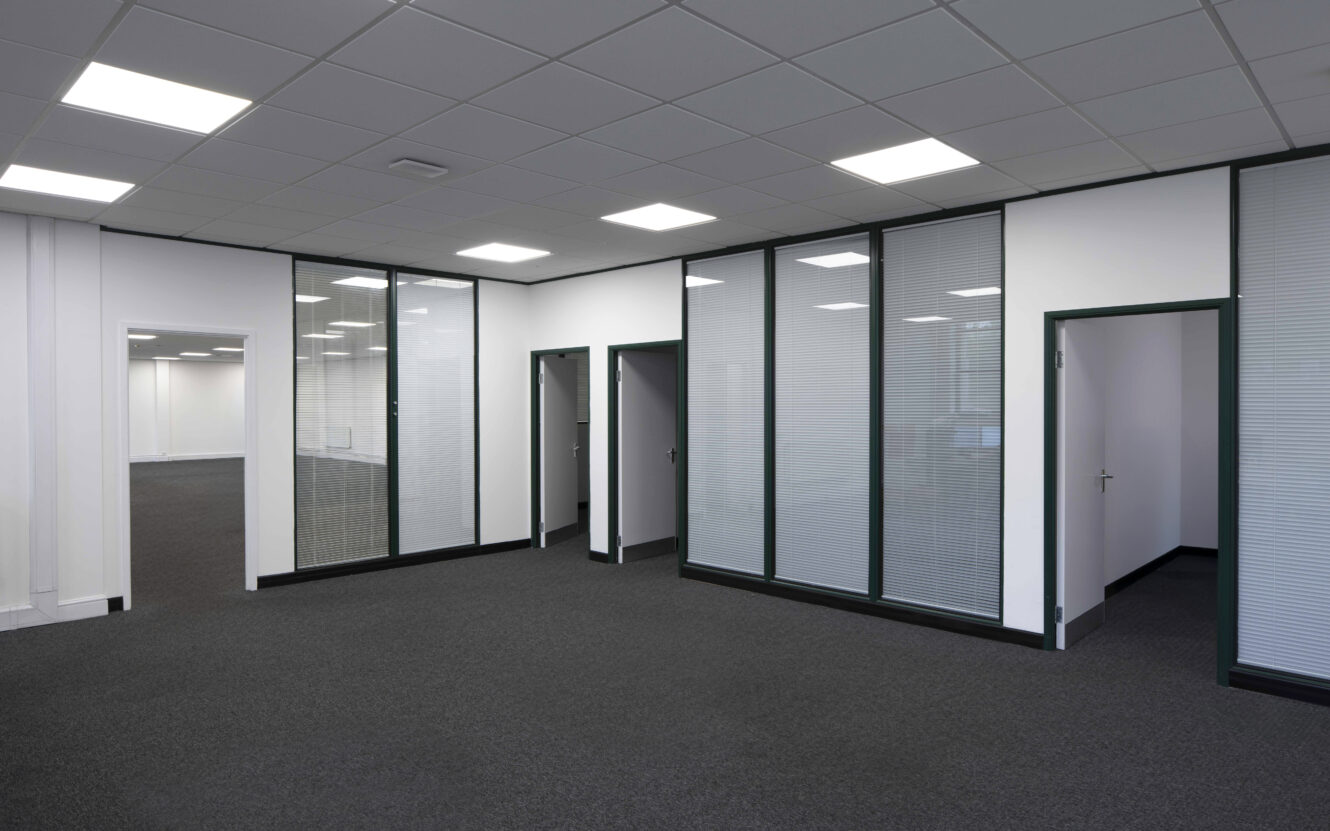 Modern office space with glass-partitioned private rooms, closed doors, white walls, and a carpeted floor under bright ceiling lights.