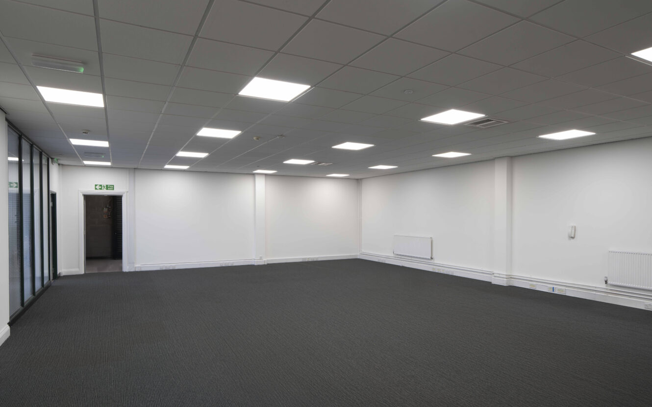 Empty office space with white walls, grey carpet, ceiling lights, radiators, and a glass wall on the left side. An open door leads to another area.