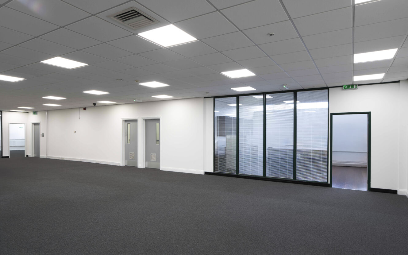 Empty modern office space with gray carpet, white walls, glass-partitioned room, ceiling lights, and closed doors.