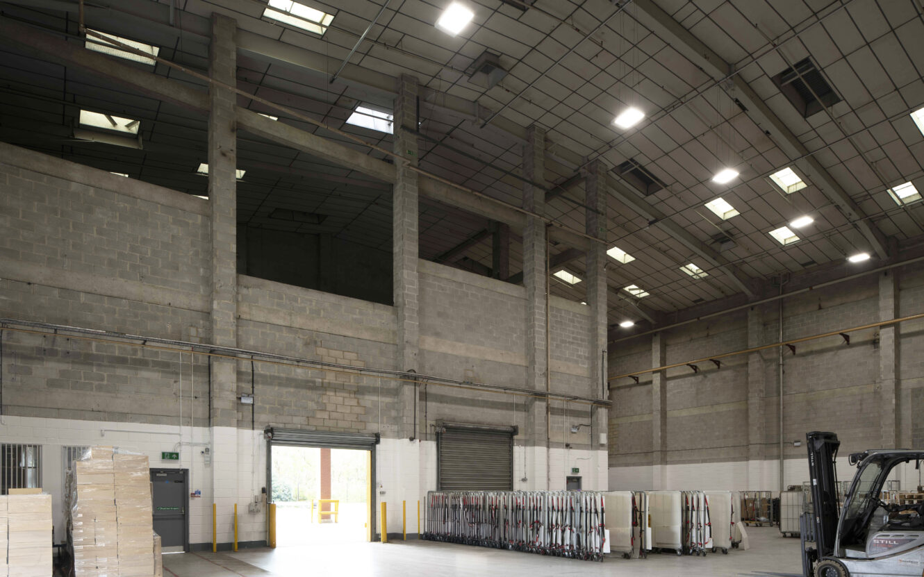 A spacious industrial warehouse interior with high ceilings, concrete walls, stacked pallets, a forklift, and overhead lighting.