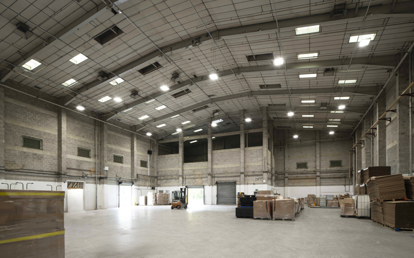 Large, empty warehouse with high ceilings, concrete floors, exposed brick walls, overhead lighting, and stacks of pallets and cardboard boxes along the side.