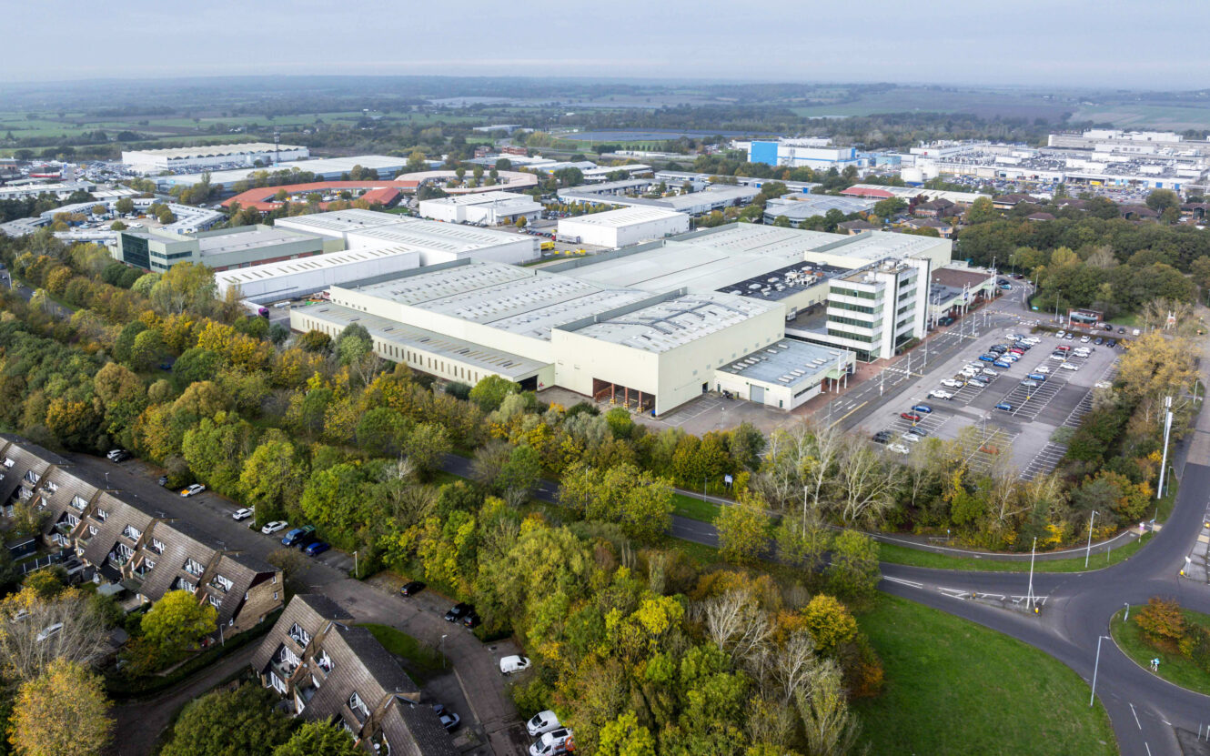 Aerial view of a large industrial complex surrounded by trees, with parking lots, nearby residential houses, and surrounding roads visible.