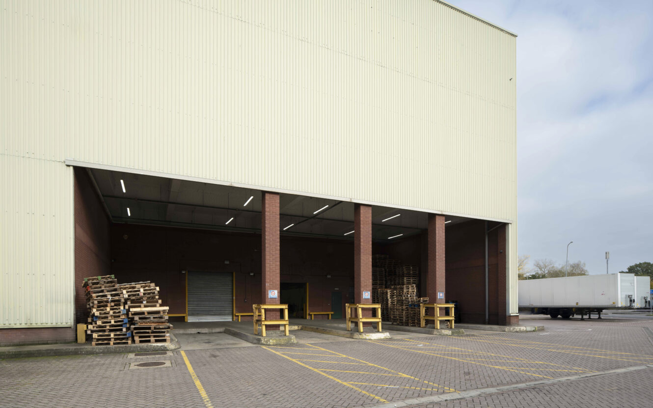 A warehouse loading dock with stacked wooden pallets, yellow safety barriers, and a parked white truck trailer in a paved lot.