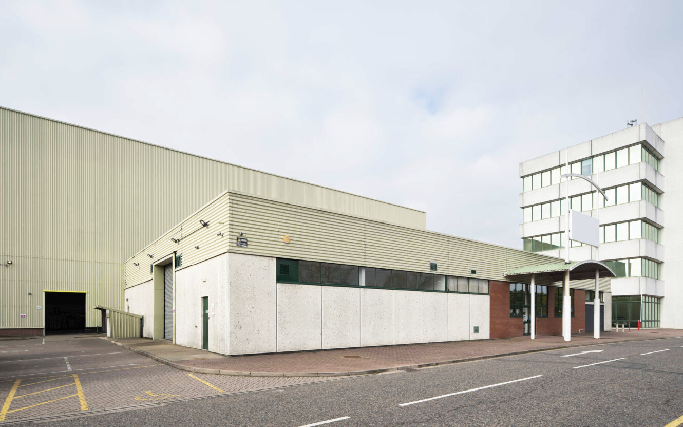 A large industrial warehouse and adjacent office building with a mostly empty parking area and overcast sky.