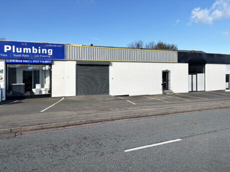 Single-story commercial building with a plumbing store and adjacent empty parking spaces, viewed from across the street on a bright day.