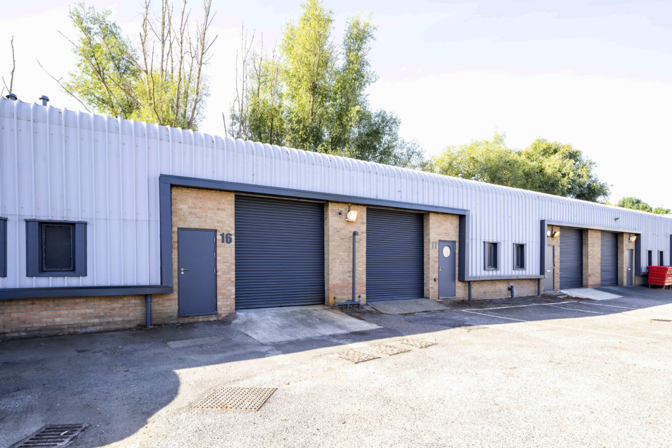 A row of industrial warehouse units with brick and metal walls, closed grey roller doors, numbered 16, and a few trees in the background.