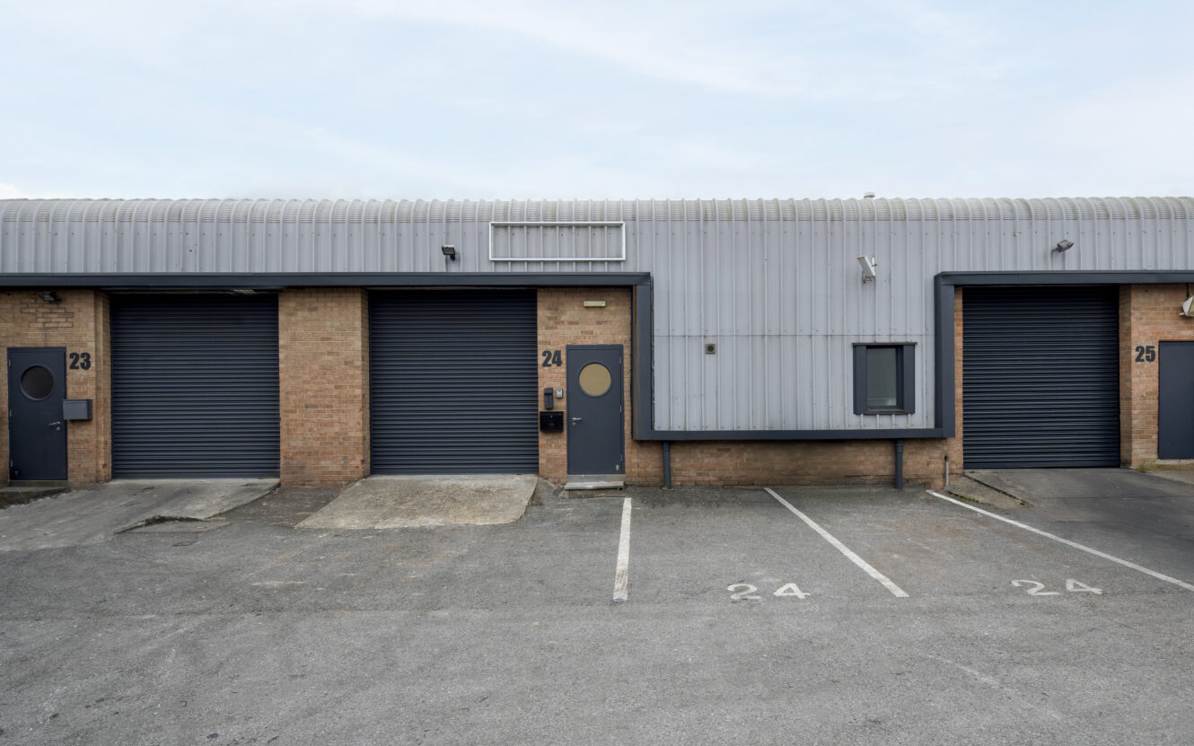 A row of industrial warehouse units with closed gray roller doors, numbered 23, 24, and 25, and empty parking spaces marked 24 and 25 in front.