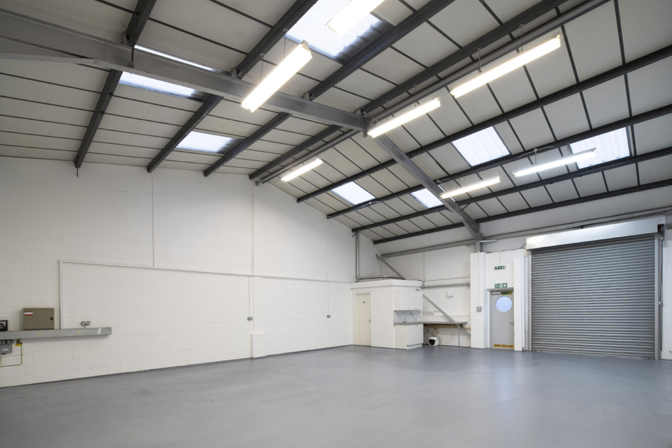 Empty industrial warehouse with white walls, gray concrete floor, metal ceiling beams, skylights, fluorescent lights, a closed roller door, and utility fixtures along one wall.