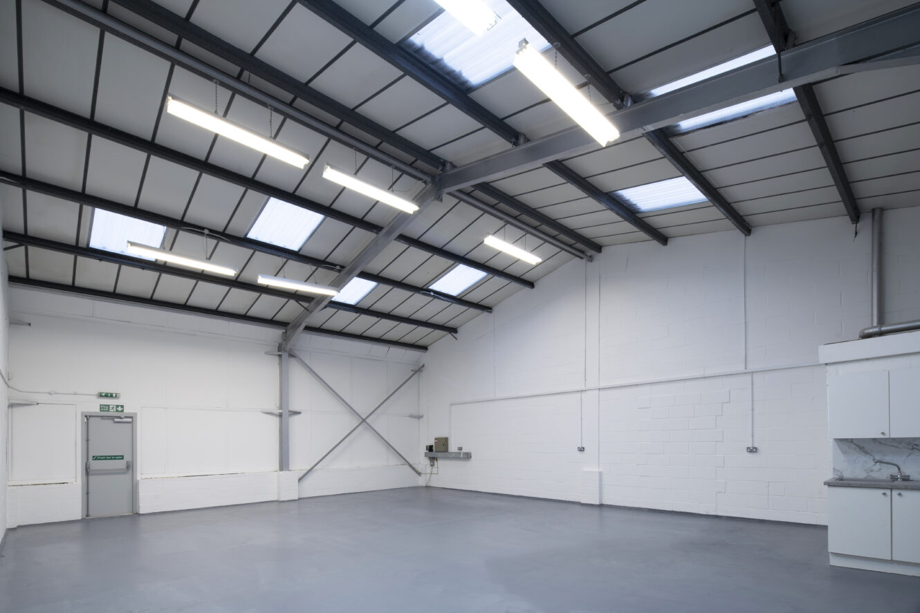 Spacious, empty warehouse interior with high ceiling, white walls, exposed beams, skylights, fluorescent lights, and a closed door on the left.