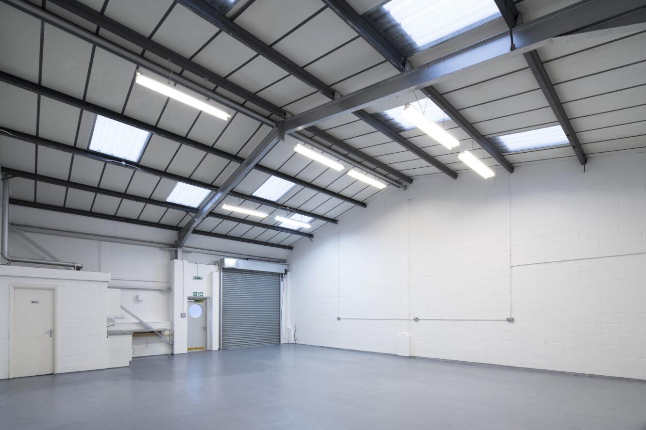 Empty industrial warehouse with gray concrete floor, white walls, metal ceiling, fluorescent lights, two doors, a roller shutter door, and skylights.