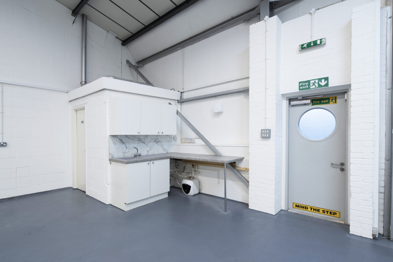 A small kitchenette with white cabinets and a marble backsplash is in the corner of an industrial space. A door with a round window and Mind the Step sign is on the right.