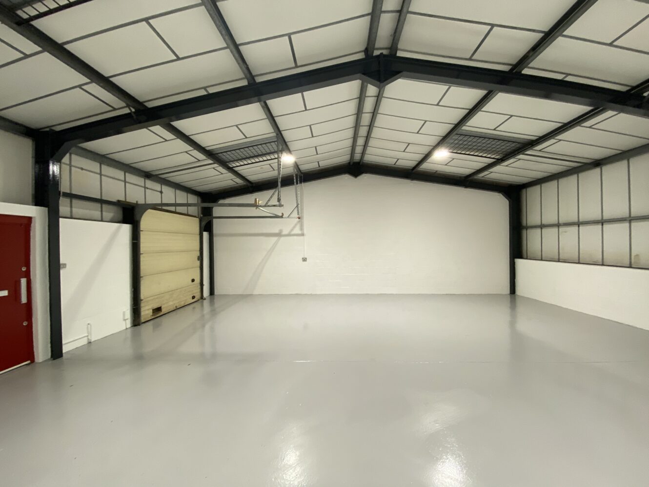 Empty industrial warehouse with white walls, a polished gray floor, high ceiling, a large garage door, and a red entry door on the left side.