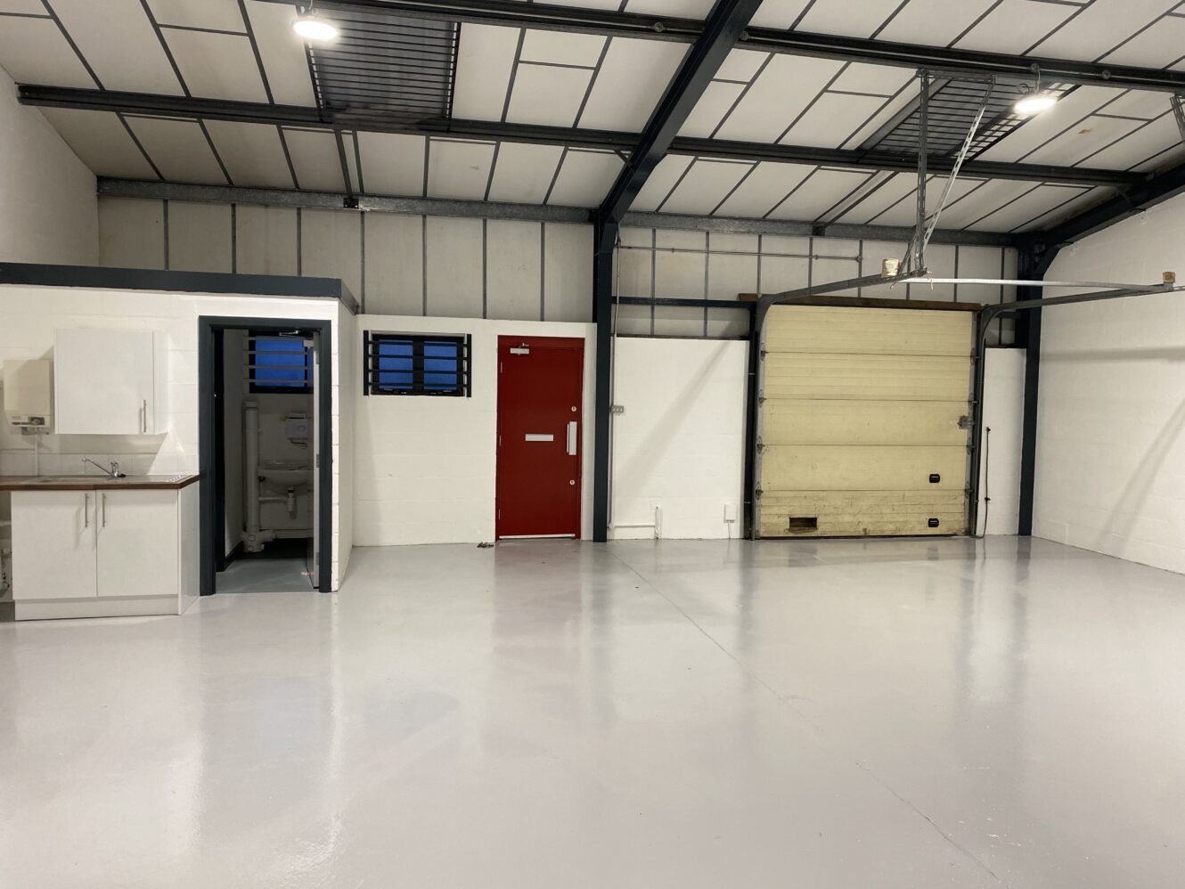 Interior of an empty industrial unit with a small kitchenette, a restroom, a red door, and a large roller shutter door. The floor is clean and gray.