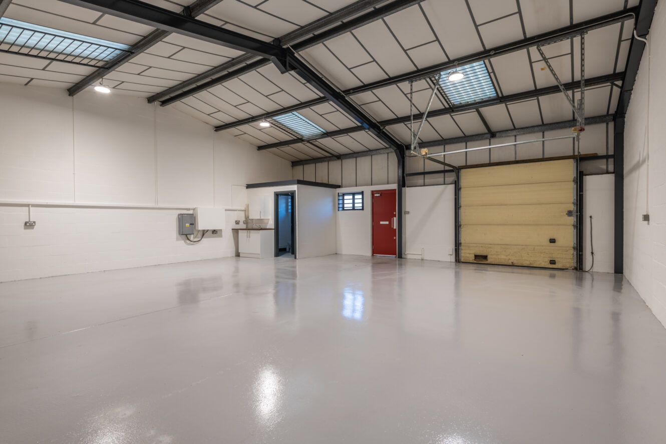 Empty industrial warehouse with a polished concrete floor, high ceiling, fluorescent lighting, a red door, small windows, and a large yellow roll-up garage door.